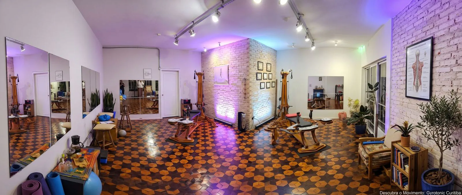 Vista panorâmica do estúdio, com equipamentos GYROTONIC, parede de tijolinho com certificados emoldurados e piso de tacos em mosaico